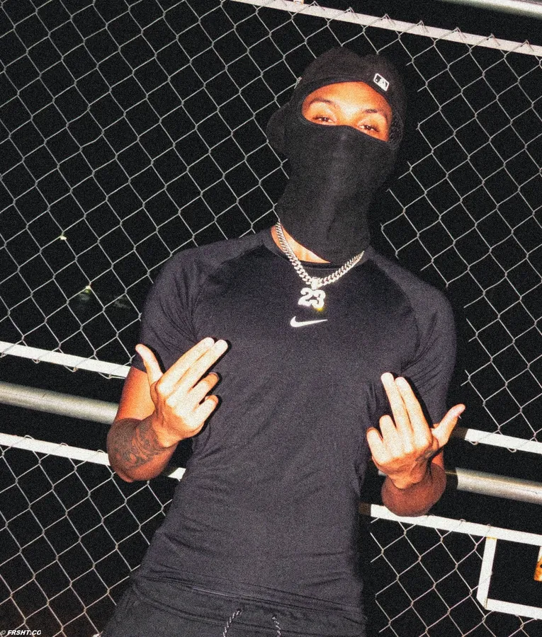 Artista de balaclava e boné, fazendo gesto com as mãos em frente a uma grade metálica