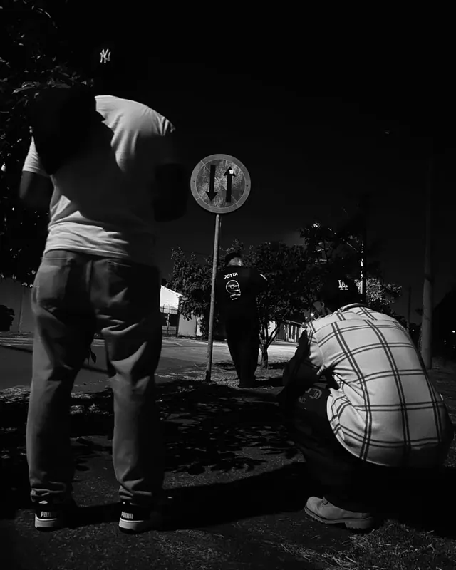 Cena noturna em preto e branco, com três pessoas ao redor de uma placa de trânsito